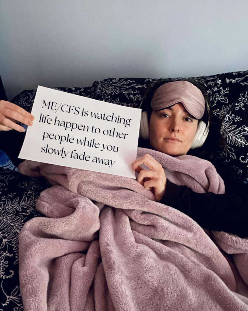 Samantha is holding a sign that reads: ME/CFS is watching life happen to other people while you slowly fade away.