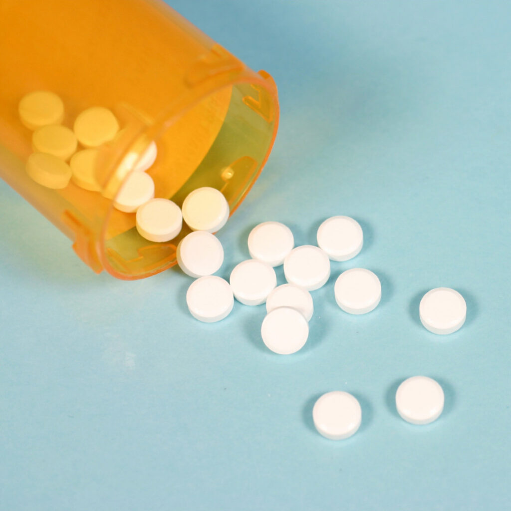 The image shows an orange prescription pill bottle tipped on its side, with small white round tablets spilling out onto a light blue surface.